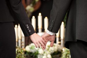 Two people holding hands at a Jewish funeral in front of candles and flowers.